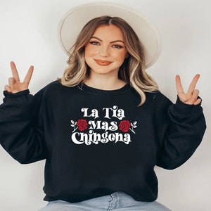 Op de afbeelding: Een zwart sweatshirt met de tekst "La Tia Mas Chingona" in wit met een rode roos afbeelding.