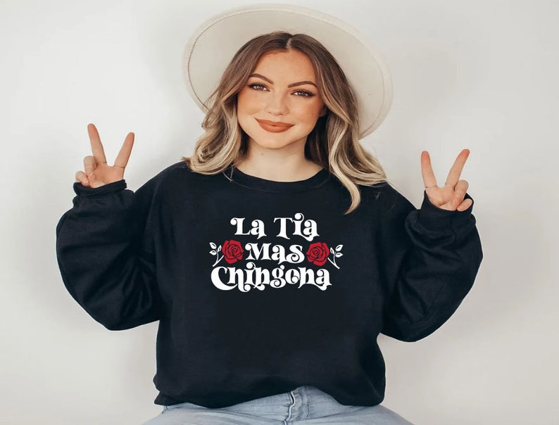Op de afbeelding: Een zwart sweatshirt met de tekst "La Tia Mas Chingona" in wit met een rode roos afbeelding.