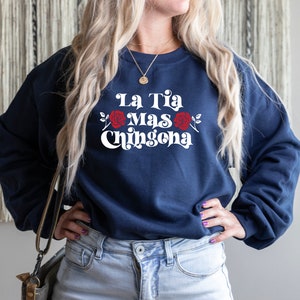Op de afbeelding: Een marineblauw sweatshirt met de tekst "La Tia Mas Chingona" in wit met rode rozen aan weerszijden van de tekst.