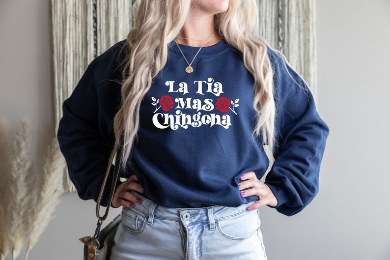 Op de afbeelding: Een marineblauw sweatshirt met de tekst "La Tia Mas Chingona" in wit met rode rozen aan weerszijden van de tekst.