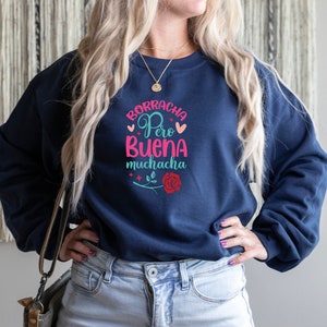May include: A navy blue sweatshirt with the text "Borracha Pero Buena Muchacha" in pink, blue, and green lettering. The text is surrounded by hearts and stars. A red rose with a green stem is below the text.