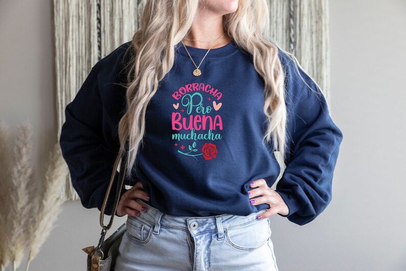 May include: A navy blue sweatshirt with the text "Borracha Pero Buena Muchacha" in pink, blue, and green lettering. The text is surrounded by hearts and stars. A red rose with a green stem is below the text.