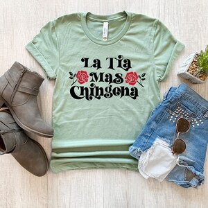 May include: A light green t-shirt with the text "La Tia Mas Chingona" in black with red roses. The shirt is laid flat on a wooden surface with a pair of brown ankle boots, a pair of blue denim shorts, and a small potted plant.