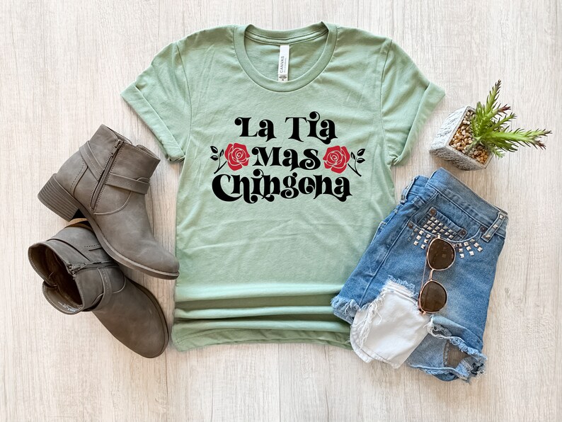 May include: A light green t-shirt with the text "La Tia Mas Chingona" in black with red roses. The shirt is laid flat on a wooden surface with a pair of brown ankle boots, a pair of blue denim shorts, and a small potted plant.