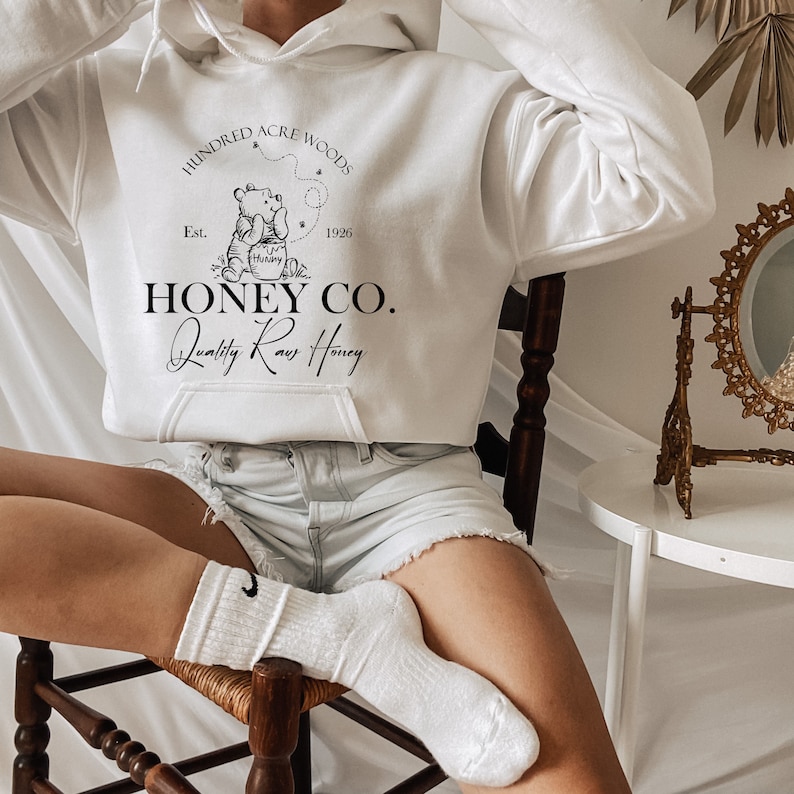 May include: White hooded sweatshirt with a black graphic design featuring Winnie the Pooh and the text "Hundred Acre Woods Est. 1926 Honey Co. Quality Raw Honey".