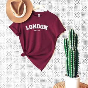 May include: A maroon t-shirt with the text "LONDON ENGLAND" in white lettering. The shirt is hanging on a white hanger against a white background with a black geometric pattern. A brown hat and a cactus in a white pot are also in the image.