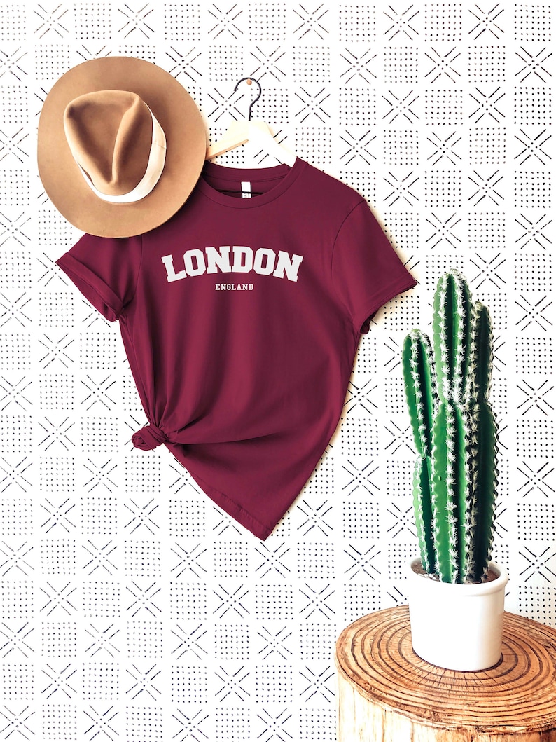 May include: A maroon t-shirt with the text "LONDON ENGLAND" in white lettering. The shirt is hanging on a white hanger against a white background with a black geometric pattern. A brown hat and a cactus in a white pot are also in the image.