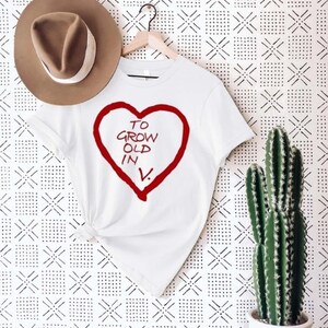 May include: White t-shirt with a red heart design and the text "TO GROW OLD IN V." A brown hat hangs above the shirt. A cactus in a white pot is on the right. The background has a black and white geometric pattern.