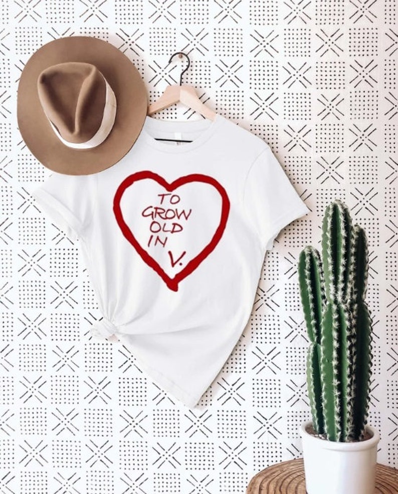 May include: White t-shirt with a red heart design and the text "TO GROW OLD IN V." A brown hat hangs above the shirt. A cactus in a white pot is on the right. The background has a black and white geometric pattern.