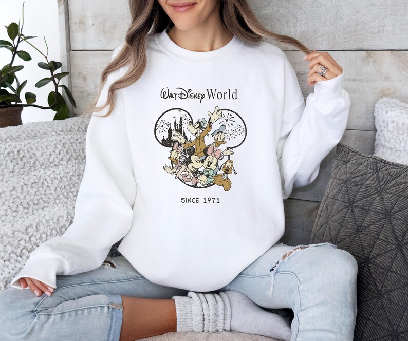 Puede incluir: Sudadera blanca con un gr&aacute;fico de Mickey Mouse y sus amigos frente al Castillo de Cenicienta. El texto "Walt Disney World" y "Since 1971" est&aacute; impreso en la sudadera.