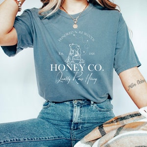 May include: A blue t-shirt with a white graphic of Winnie the Pooh holding a jar of honey. The graphic includes the text "Hundred Acre Woods", "Est. 1926", "Hunny", "Honey Co.", and "Quality Raw Honey".
