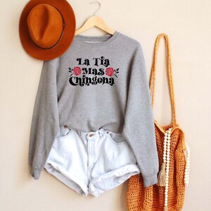Op de afbeelding: Grijs sweatshirt met de tekst "La Tia Mas Chingona" in zwart met rode rozen. Het sweatshirt hangt aan een hanger met een bruine hoed en een gehaakte tas met witte kralen.