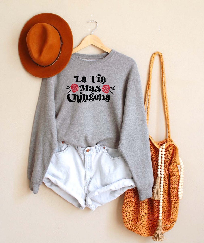 Op de afbeelding: Grijs sweatshirt met de tekst "La Tia Mas Chingona" in zwart met rode rozen. Het sweatshirt hangt aan een hanger met een bruine hoed en een gehaakte tas met witte kralen.
