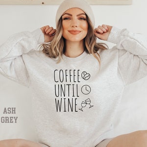 May include: A light gray sweatshirt with the text "Coffee Until Wine" in black lettering. The text is accompanied by a coffee bean, a clock, and two wine glasses.