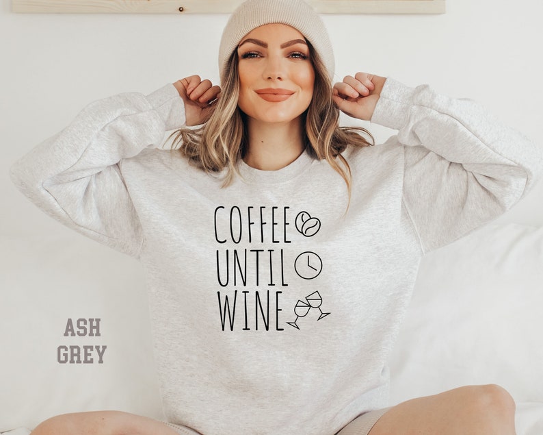 May include: A light gray sweatshirt with the text "Coffee Until Wine" in black lettering. The text is accompanied by a coffee bean, a clock, and two wine glasses.