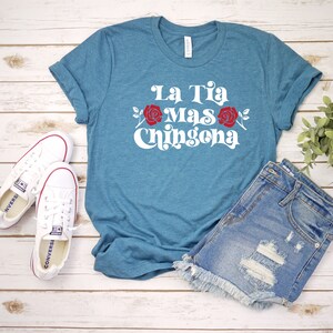 May include: A teal blue t-shirt with the text "La Tia Mas Chingona" in white with red roses on either side of the text. The shirt is laid out on a wooden surface with a pair of white sneakers and denim shorts.