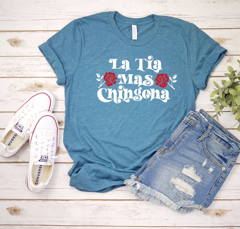 May include: A teal blue t-shirt with the text "La Tia Mas Chingona" in white with red roses on either side of the text. The shirt is laid out on a wooden surface with a pair of white sneakers and denim shorts.