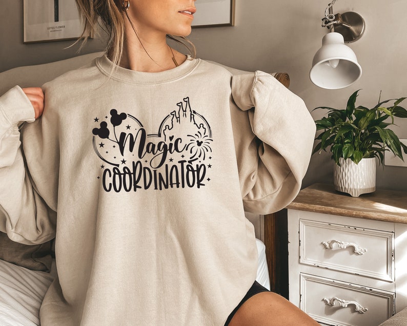 May include: A beige sweatshirt with a black graphic of Mickey Mouse ears, a castle, and fireworks. The text "Magic Coordinator" is printed below the graphic.