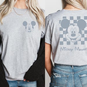 Pu&ograve; includere: Una maglietta grigia con un motivo a scacchi bianco e nero e un'immagine grigia di Topolino sul retro. Il testo "Mickey Mouse" &egrave; stampato sotto l'immagine.