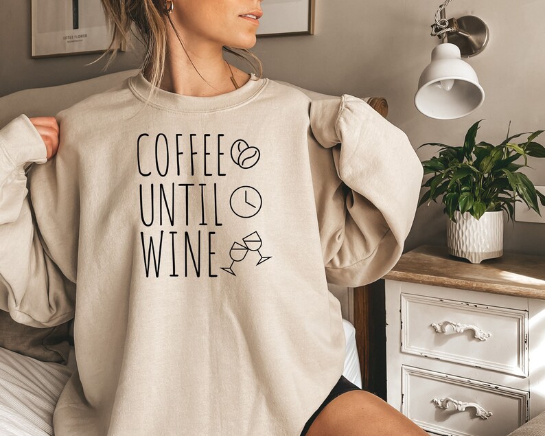 May include: A beige sweatshirt with the text "Coffee Until Wine" in black lettering. The text is accompanied by a coffee bean icon, a clock icon, and two wine glass icons.