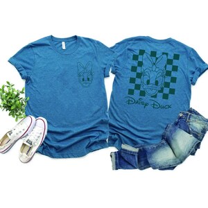 Pu&ograve; includere: Due magliette blu con un contorno verde di Daisy Duck. Una maglietta ha un motivo a scacchi e il testo "Daisy Duck". L'altra maglietta ha un'immagine pi&ugrave; piccola di Daisy Duck.