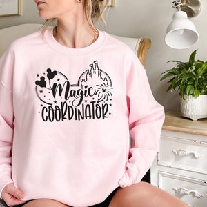 May include: Pink sweatshirt with a black graphic of Mickey Mouse ears, a castle, fireworks, and the text "Magic Coordinator".