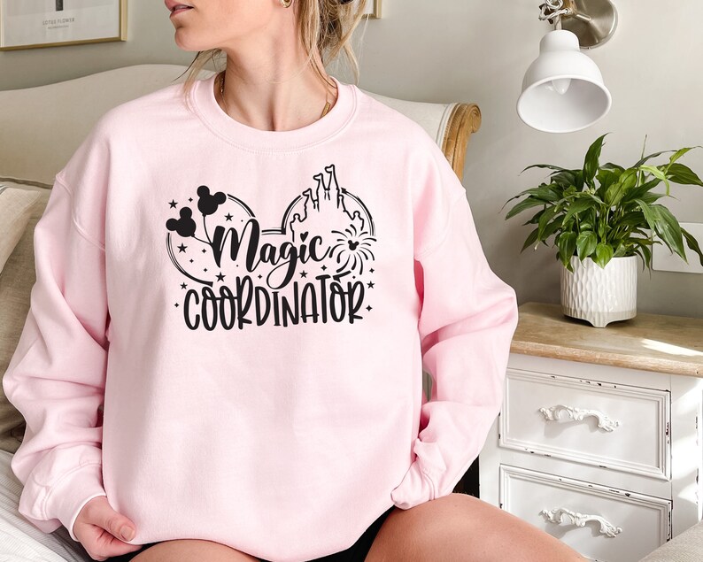 May include: Pink sweatshirt with a black graphic of Mickey Mouse ears, a castle, fireworks, and the text "Magic Coordinator".