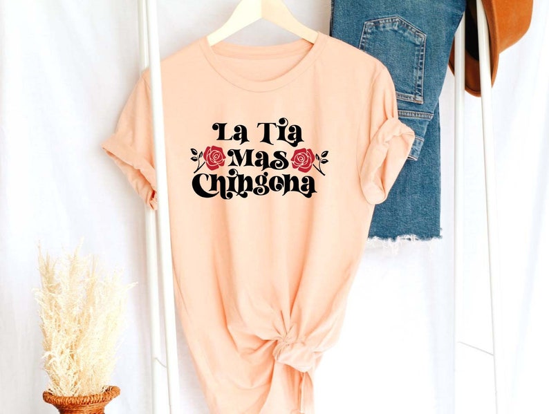 May include: Peach colored t-shirt with black text that reads "La Tia Mas Chingona" with a red rose graphic.