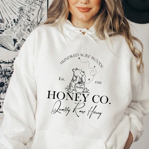 May include: White hoodie with a black graphic design featuring Winnie the Pooh holding a jar of honey. The graphic includes the text "Hundred Acre Woods", "Est. 1926", "Honey Co.", and "Quality Raw Honey".