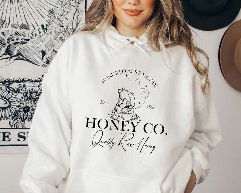 May include: White hoodie with a black graphic design featuring Winnie the Pooh holding a jar of honey. The graphic includes the text "Hundred Acre Woods", "Est. 1926", "Honey Co.", and "Quality Raw Honey".