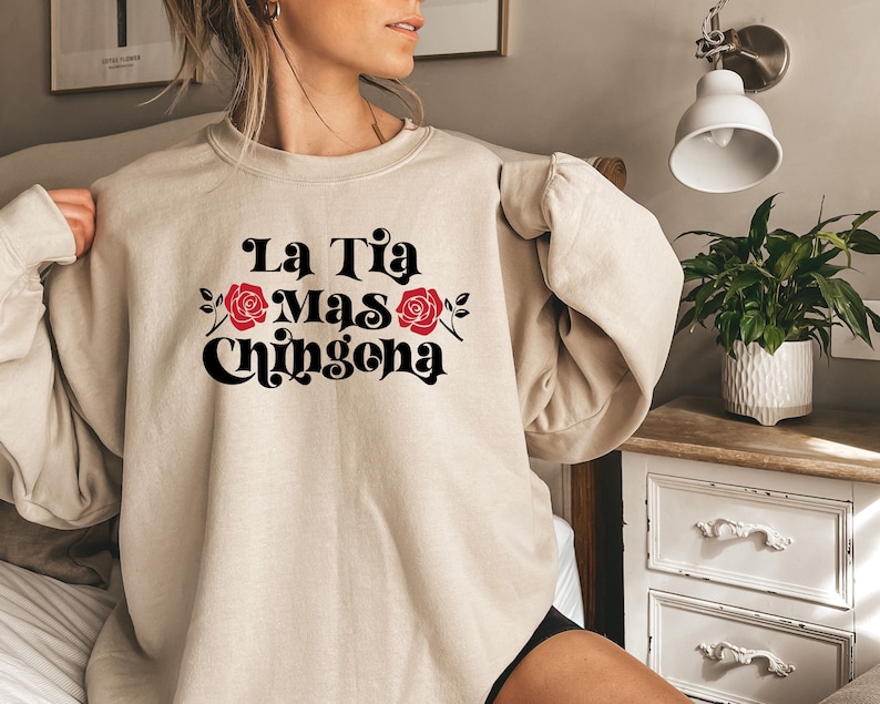Op de afbeelding: Een beige sweatshirt met de tekst "La Tia Mas Chingona" in zwart met rode rozen aan weerszijden van de tekst.
