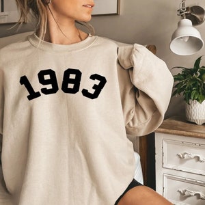 Pode incluir: Uma camisa de moletom bege com o ano 1983 impresso em preto na frente.