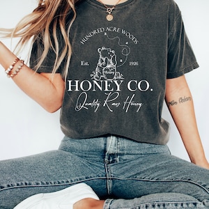 May include: A dark gray t-shirt with a white graphic of Winnie the Pooh holding a jar of honey. The graphic includes the text "Hundred Acre Woods", "Est. 1926", "Honey Co.", and "Quality Raw Honey".