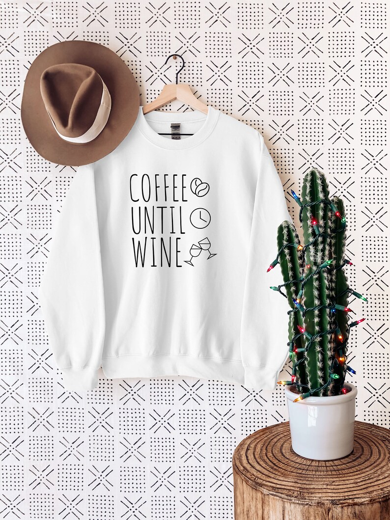 May include: White sweatshirt with the text "Coffee Until Wine" in black lettering. A coffee bean, clock, and wine glass icons are included in the design.