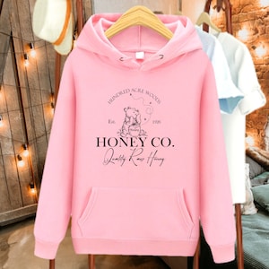 May include: Pink hoodie with a black graphic of two bears with a honey pot and the text "Hundred Acre Woods Est. 1926 Honey Co. Quality Raw Honey".