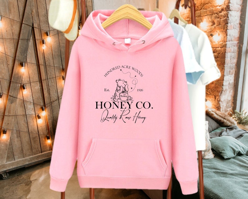 May include: Pink hoodie with a black graphic of two bears with a honey pot and the text "Hundred Acre Woods Est. 1926 Honey Co. Quality Raw Honey".