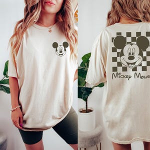 K&ouml;nnte beinhalten: Ein cremefarbenes T-Shirt mit einem schwarz-wei&szlig; karierten Muster auf der R&uuml;ckseite. Das Muster zeigt einen Mickey-Maus-Kopf aus einem Cartoon mit dem Text "Mickey Mouse" darunter. Die Vorderseite des Shirts hat einen kleinen Mickey-Maus-Kopf aus einem Cartoon in der Mitte.