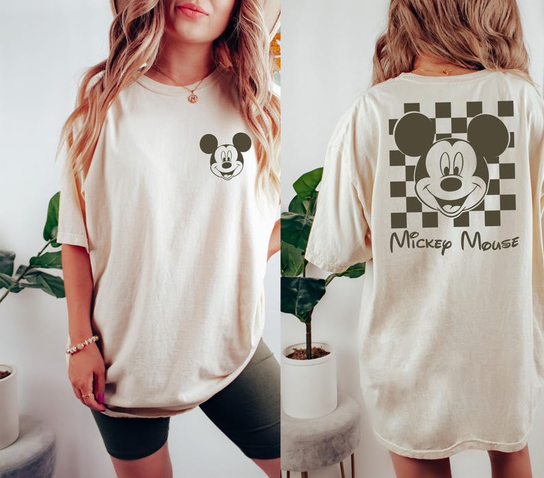 K&ouml;nnte beinhalten: Ein cremefarbenes T-Shirt mit einem schwarz-wei&szlig; karierten Muster auf der R&uuml;ckseite. Das Muster zeigt einen Mickey-Maus-Kopf aus einem Cartoon mit dem Text "Mickey Mouse" darunter. Die Vorderseite des Shirts hat einen kleinen Mickey-Maus-Kopf aus einem Cartoon in der Mitte.