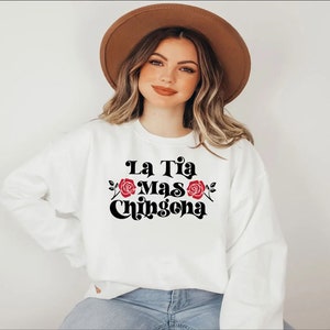 Op de afbeelding: Wit sweatshirt met zwarte tekst die "La Tia Mas Chingona" luidt met twee rode rozen aan weerszijden van de tekst.