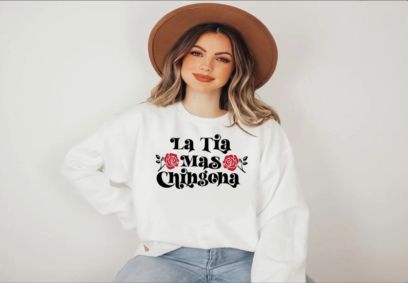 Op de afbeelding: Wit sweatshirt met zwarte tekst die "La Tia Mas Chingona" luidt met twee rode rozen aan weerszijden van de tekst.