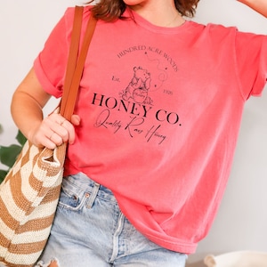 May include: A pink t-shirt with a black graphic design featuring a cartoon bear holding a jar of honey. The graphic design includes the text "Hundred Acre Woods", "Est. 1926", "Honey Co.", and "Quality Raw Honey".