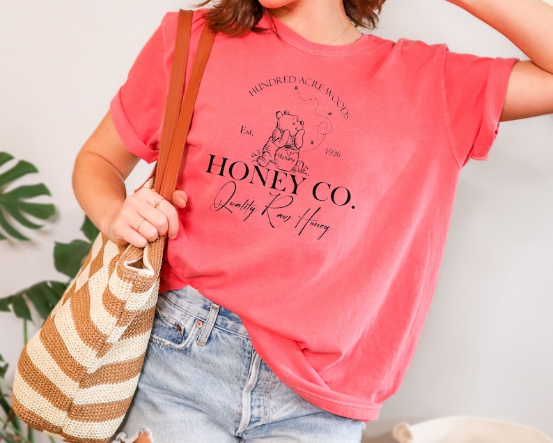 May include: A pink t-shirt with a black graphic design featuring a cartoon bear holding a jar of honey. The graphic design includes the text "Hundred Acre Woods", "Est. 1926", "Honey Co.", and "Quality Raw Honey".