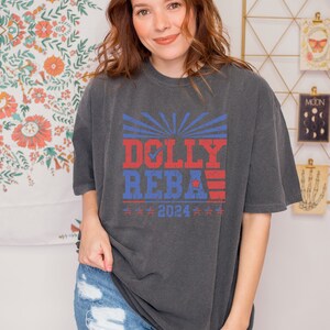 May include: A dark gray t-shirt with a red, white, and blue graphic print that says "DOLLY REBA 2024".
