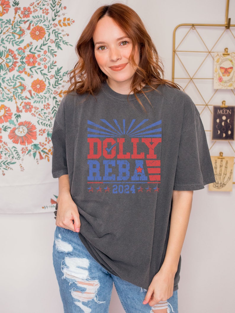 May include: A dark gray t-shirt with a red, white, and blue graphic print that says "DOLLY REBA 2024".