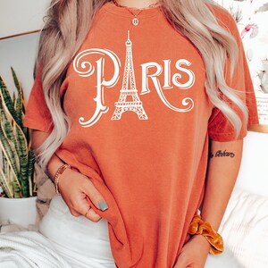 May include: A burnt orange t-shirt with a white graphic of the Eiffel Tower and the word "PARIS" in a stylized font.