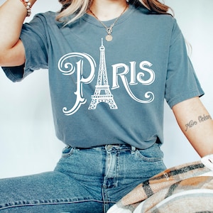 May include: A woman wearing a blue t-shirt with a white graphic of the Eiffel Tower and the word "PARIS" in a stylized font. She is also wearing blue jeans and a gold necklace.