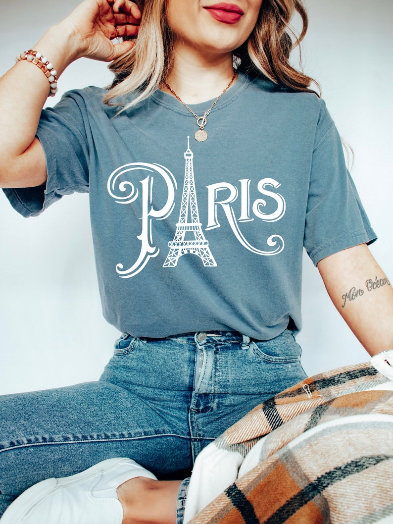 May include: A woman wearing a blue t-shirt with a white graphic of the Eiffel Tower and the word "PARIS" in a stylized font. She is also wearing blue jeans and a gold necklace.