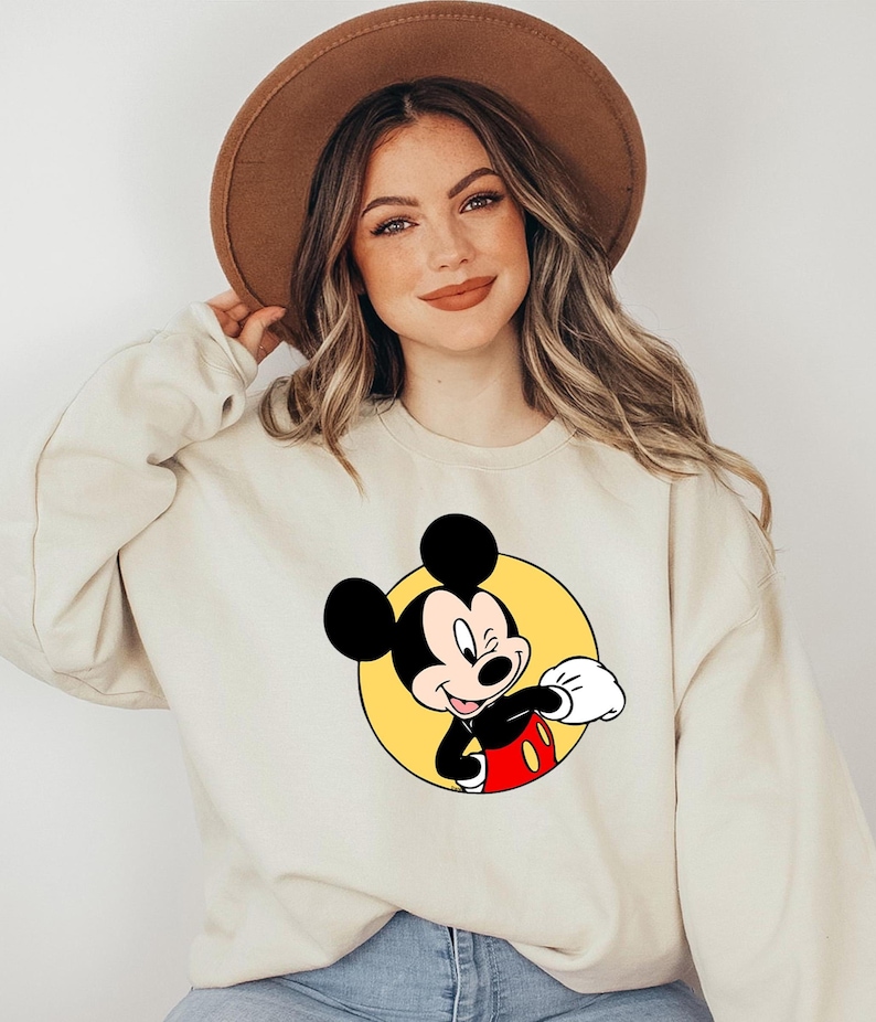 May include: A cream colored sweatshirt with a cartoon image of Mickey Mouse wearing red shorts and a yellow circle around him.