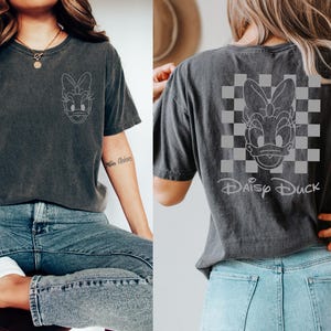 K&ouml;nnte beinhalten: Ein dunkelgraues T-Shirt mit einem wei&szlig;en Umriss von Daisy Duck auf der Vorderseite und einem wei&szlig;en Umriss von Daisy Duck auf einem karierten Hintergrund auf der R&uuml;ckseite mit dem Text "Daisy Duck" darunter.