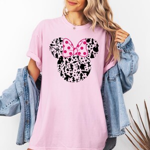 May include: A pink t-shirt with a black silhouette of Mickey Mouse's head made up of various Disney characters. The bow is pink with white polka dots.